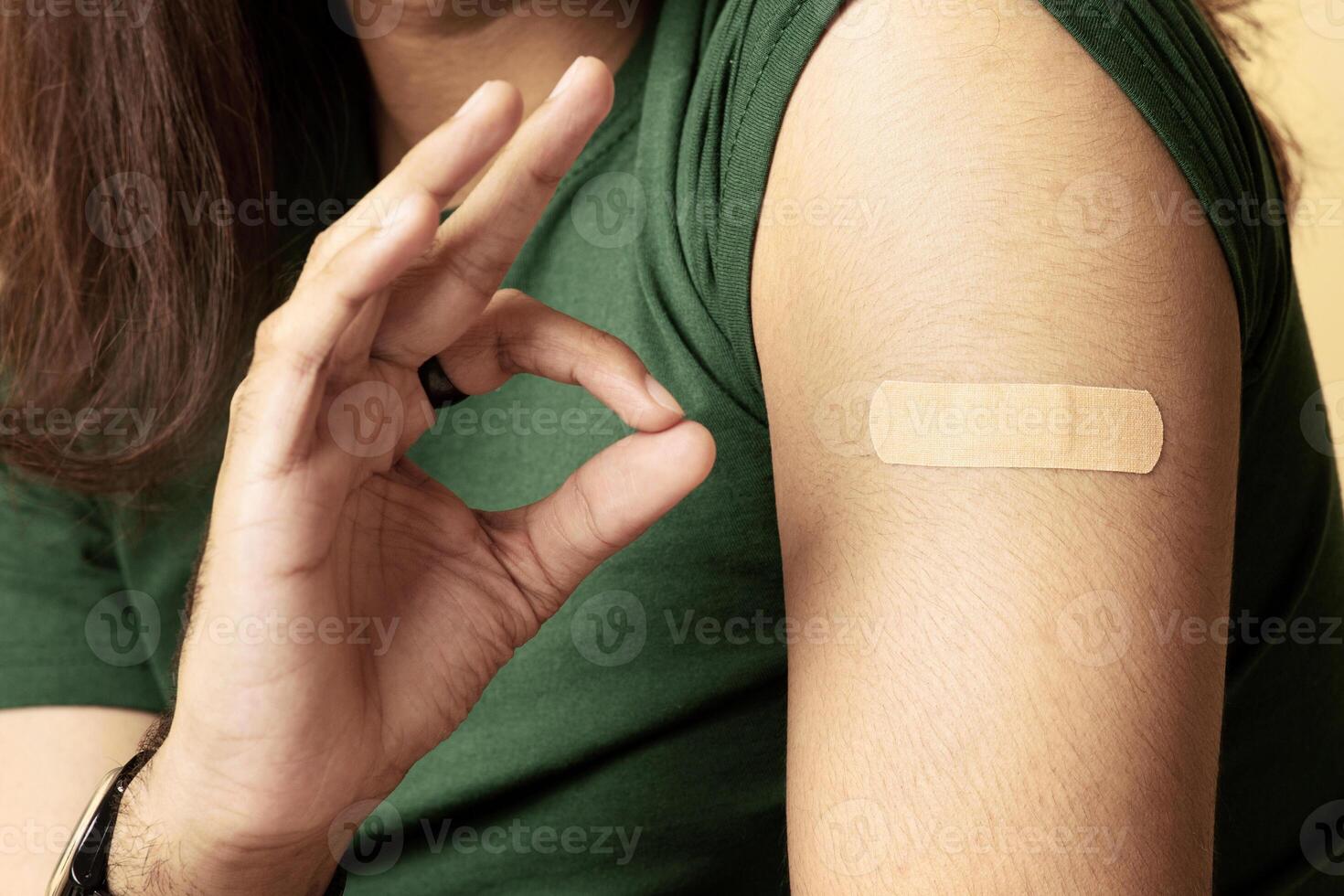 Unrecognizable man showing shoulder with medical band and okay gesture, got vaccinated against coronavirus, closeup. Cropped of middle-eastern guy recommending safety vaccination while pandemic photo