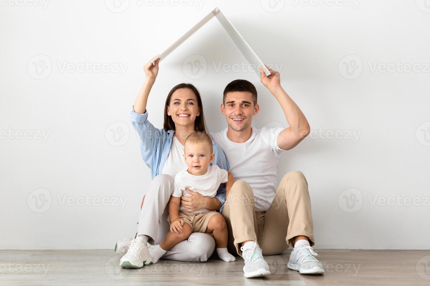 Real Estate Concept. Happy Family With Little Toddler Child Holding Symbolic Cardboard Roof Above Head, Smiling Young Spouses And Their Infant Son Dreaming About Moving To New Home, Copy Space photo
