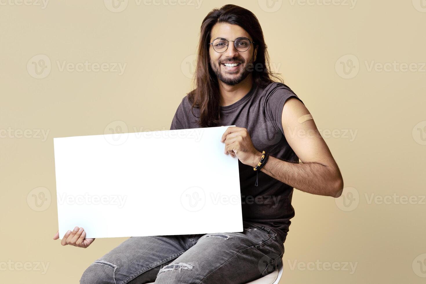 Handsome middle-eastern guy with long hair and glasses got vaccinated against COVID-19, holding horizontal white blank board, sitting on chair over beige studio background, copy space, mockup photo