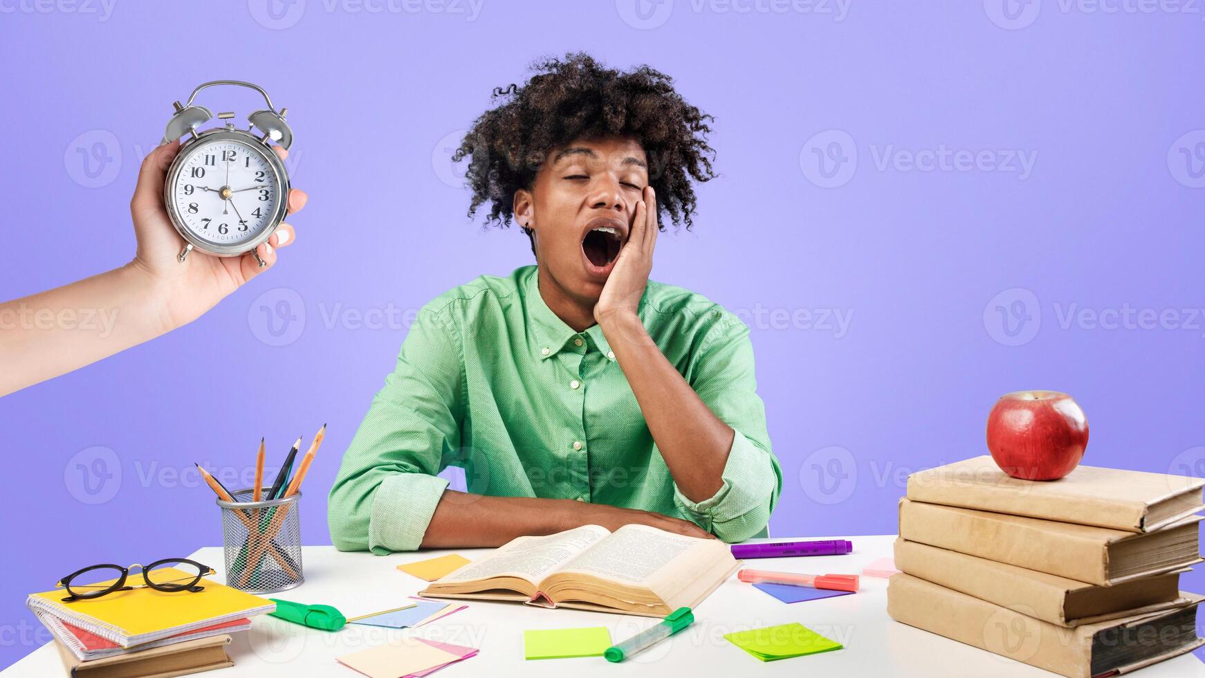 Tired african american male student yawning, sitting at desk with lot of books and preparing for exams against blue studio background. Hand with alarm clock. Deadline and overworking concept. photo
