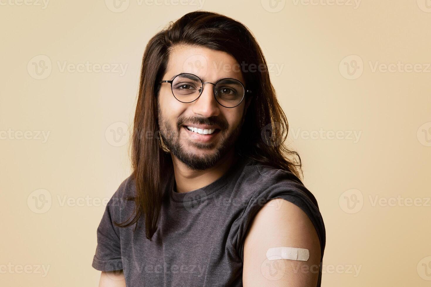 Closeup of handsome long-haired arab guy wearing glasses posing after vaccination against COVID-19, showing adhesive medical band on his shoulder and smiling at camera, studio background, copy space photo