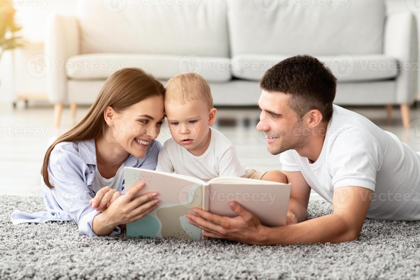 Early Education For Kids. Parents Reading Book To Their Cute Toddler Son At Home, Infant Child Looking With Interest, Happy Millennial Family Relaxing Together On Floor In Living Room, Free Space photo
