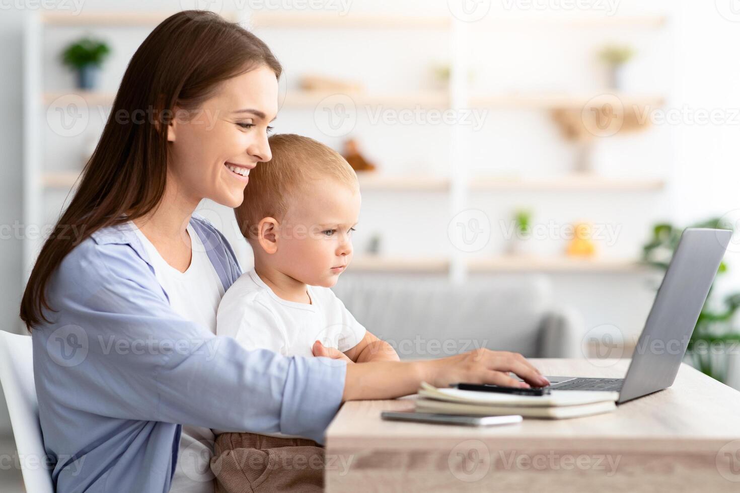 Freelance And Maternity. Young Happy Lady Working Remotely On Laptop At Home And Taking Care About Baby, Millennial Mother Sitting At Desk With Infant Son And Using Computer, Side View, Copy Space photo