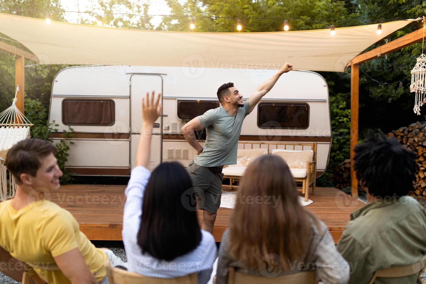 Diverse young friends playing charades near camper van, trying to guess word during outdoor party. Millennial people entertaining themselves with guessing game, having fun camping trip photo
