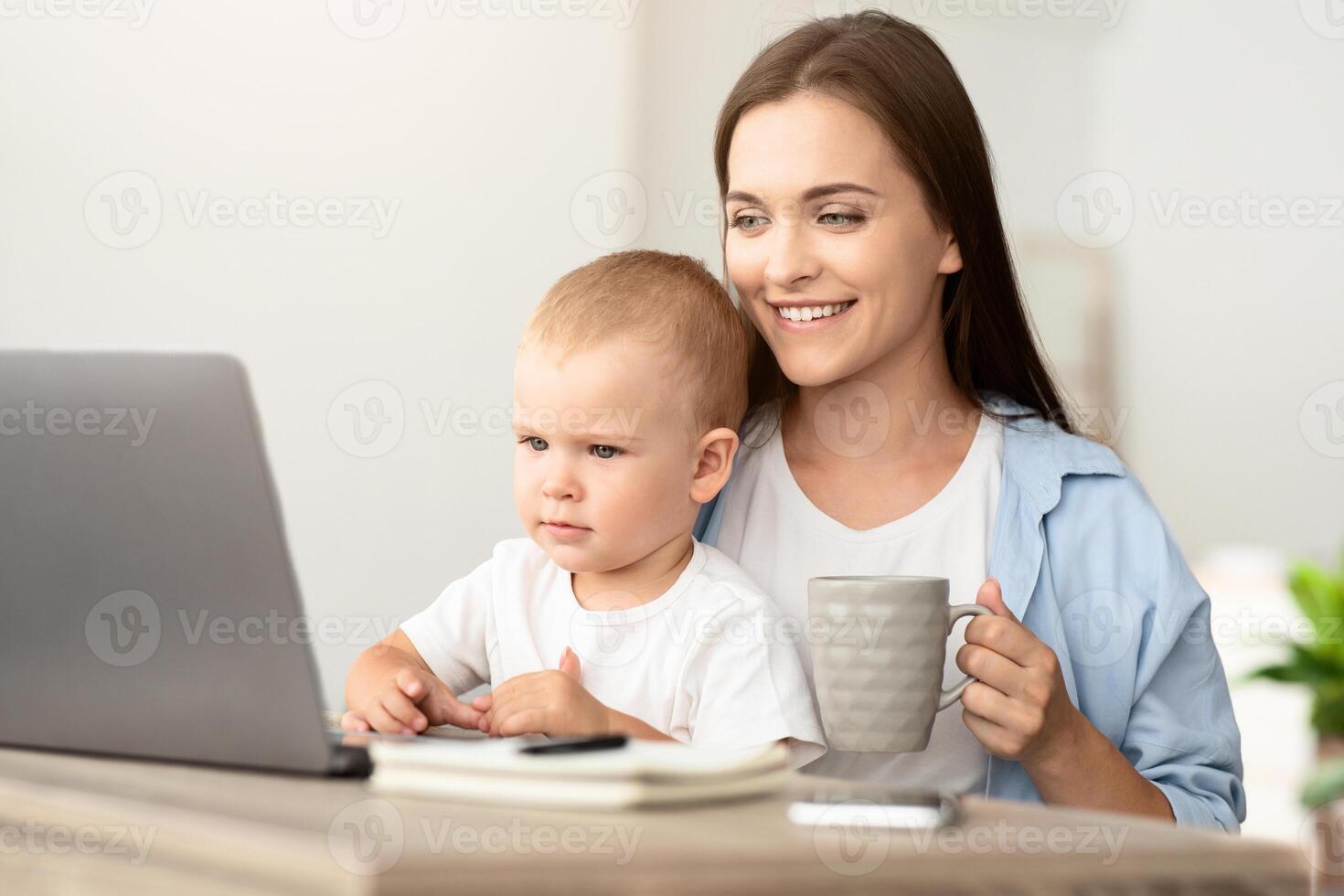 Technology And Childcare. Mother Drinking Tea While Baby Watching Cartoons On Laptop, Happy Young Mom With Toddler Son On Hands Relaxing At Home With Computer, Closeup Shot With Free Space photo