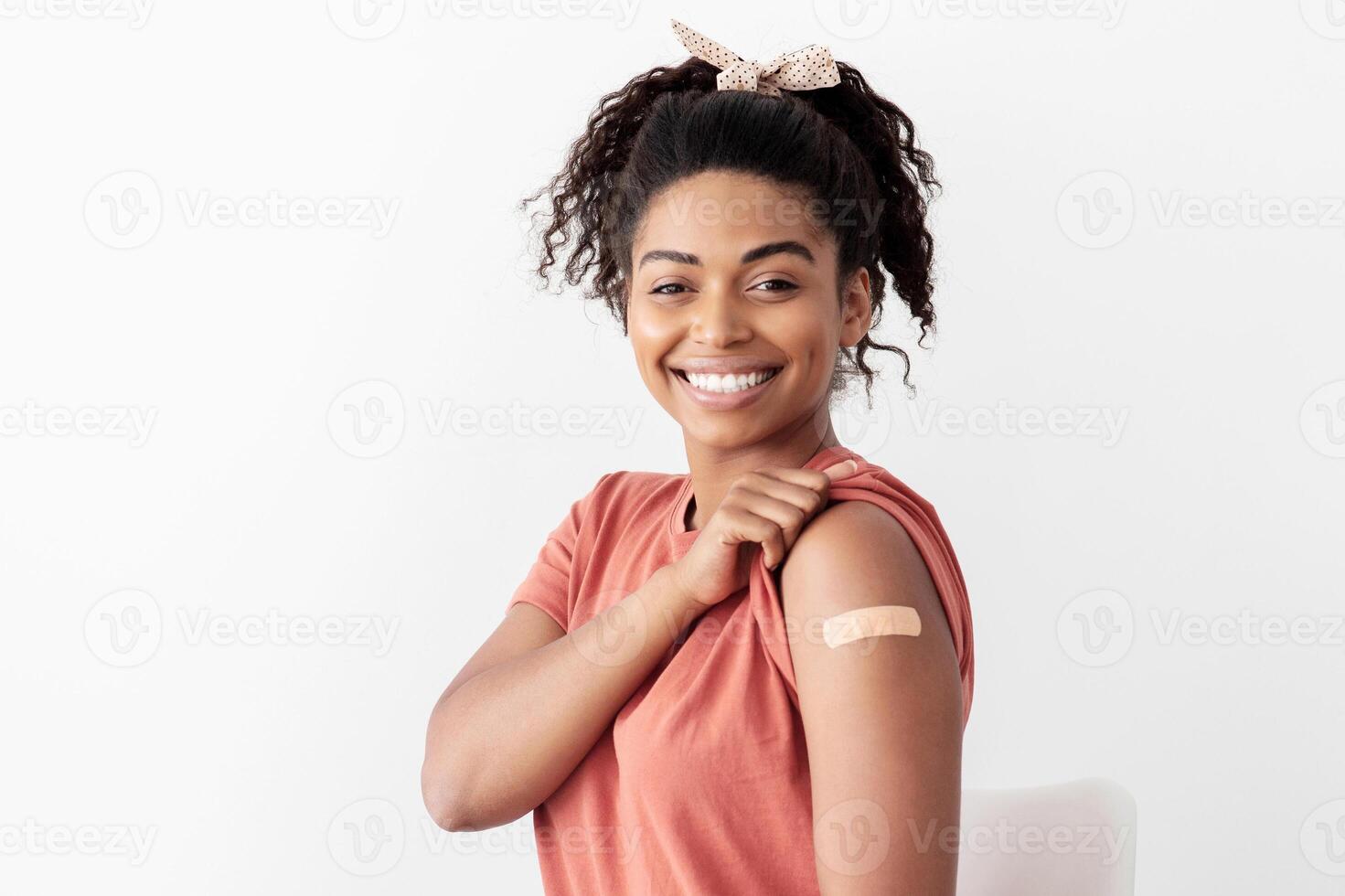 Young african american woman got vaccinated against coronavirus, sitting on chair at clinic, showing medical band on her shoulder, smiling at camera, copy space. Immunization concept photo