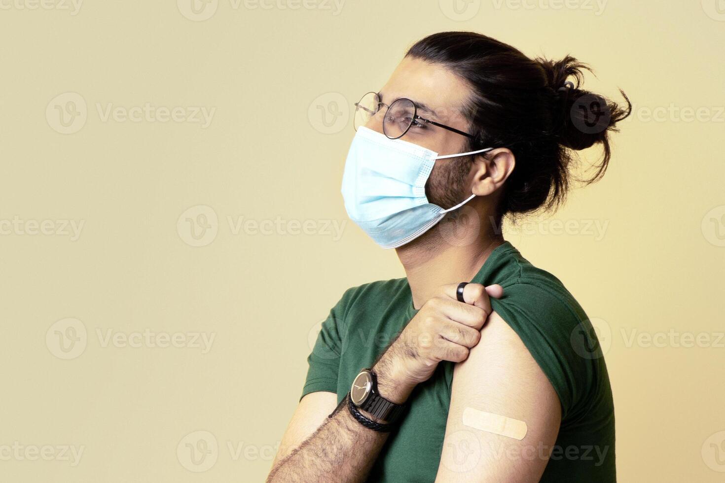 Long-haired indian man in protective face mask got vaccinated against COVID-19, showing shoulder with plaster on, sitting on chair over beige studio background, looking at copy space, panorama photo