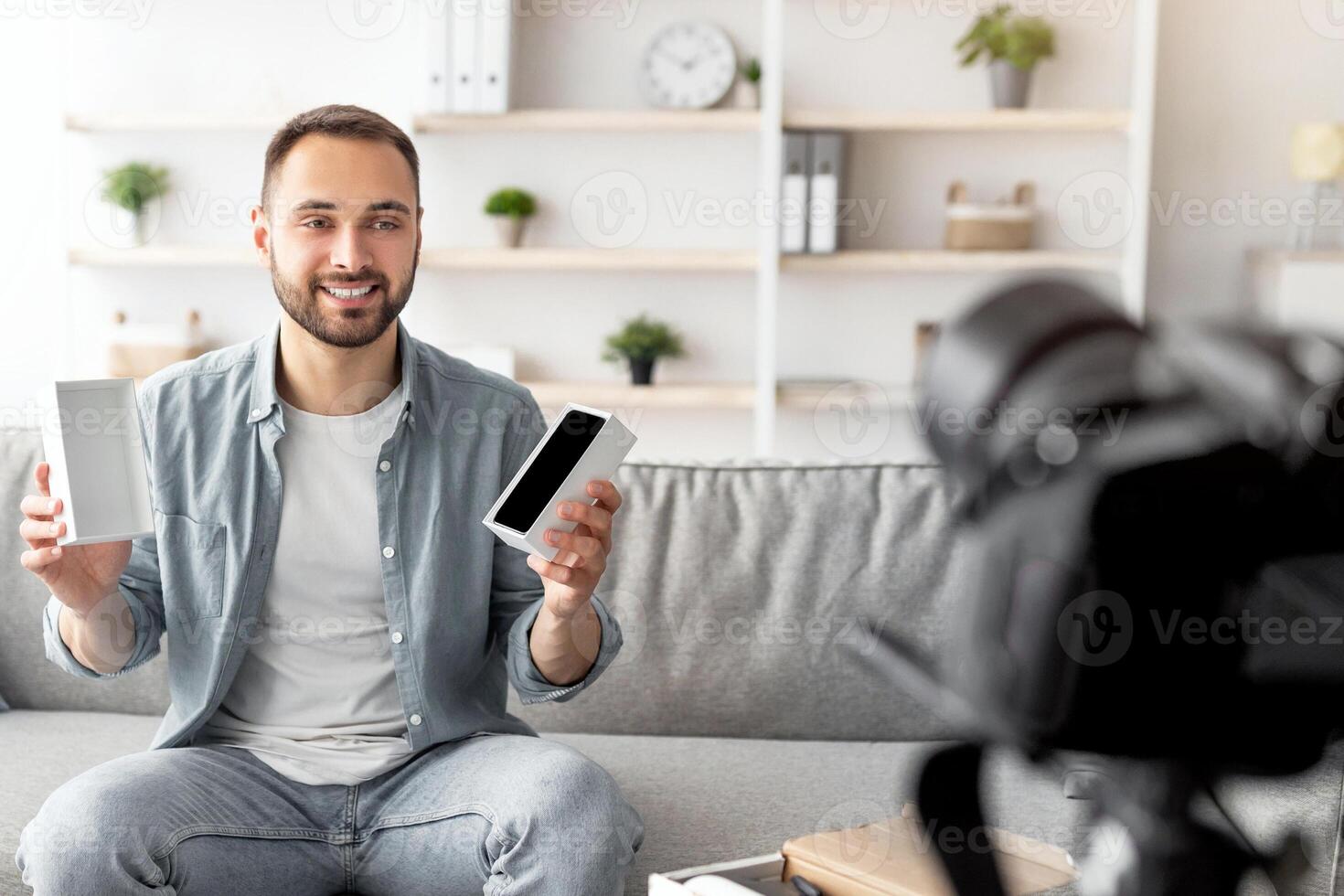 Millennial male influencer shooting review of modern smartphone, using dslr camera at home. Young content maker promoting new gadget model, filming for social media photo