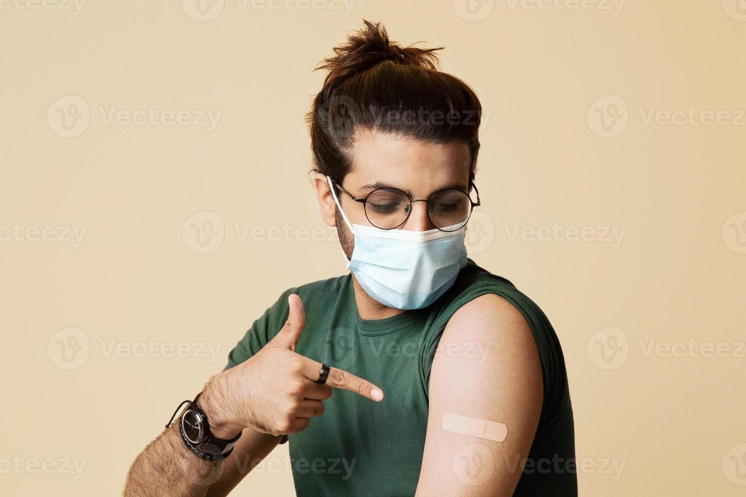 Long-haired arab guy in face mask sitting on chair over beige studio background, pointing at plaster on his shoulder, young middle-eastern man got vaccinated against COVID-19, copy space photo