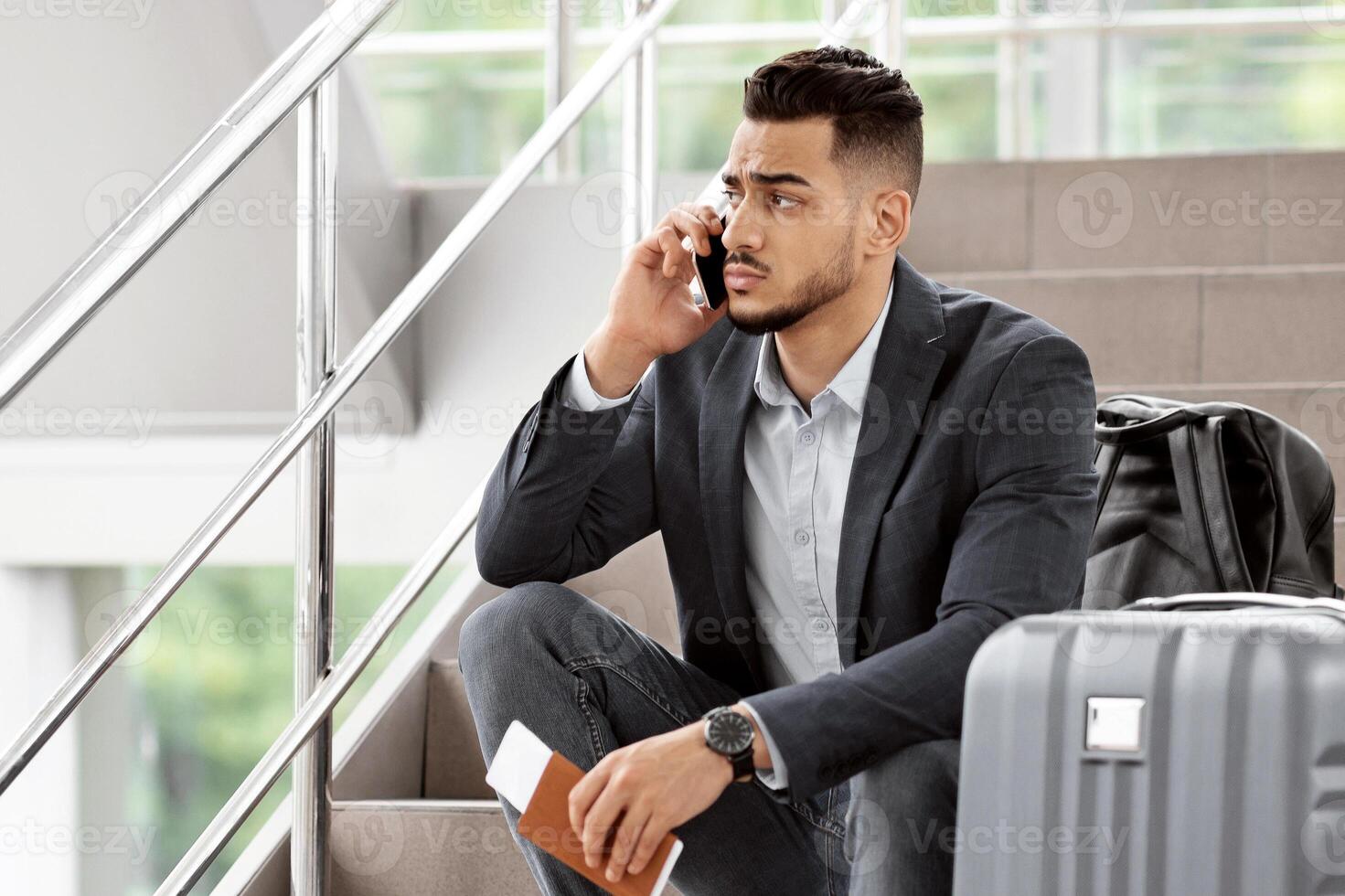 Worried Middle Eastern Man Talking On Cellphone While Sitting With Luggage On Stairs At Airport Terminal, Concerned Arab Traveller Talking To Customer Service, Changing Tickets Online, Free Space photo