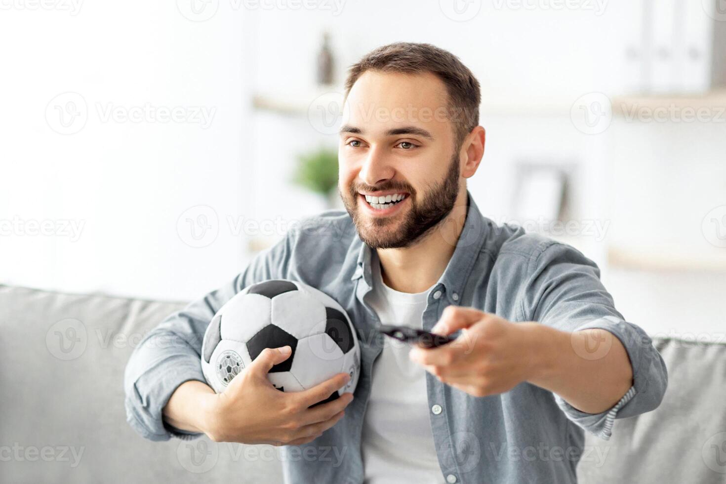 Happy young guy watching soccer game on TV, holding remote control and ball at home. Sports fan sitting on couch, enjoying television game match, cheering for his favorite football team photo