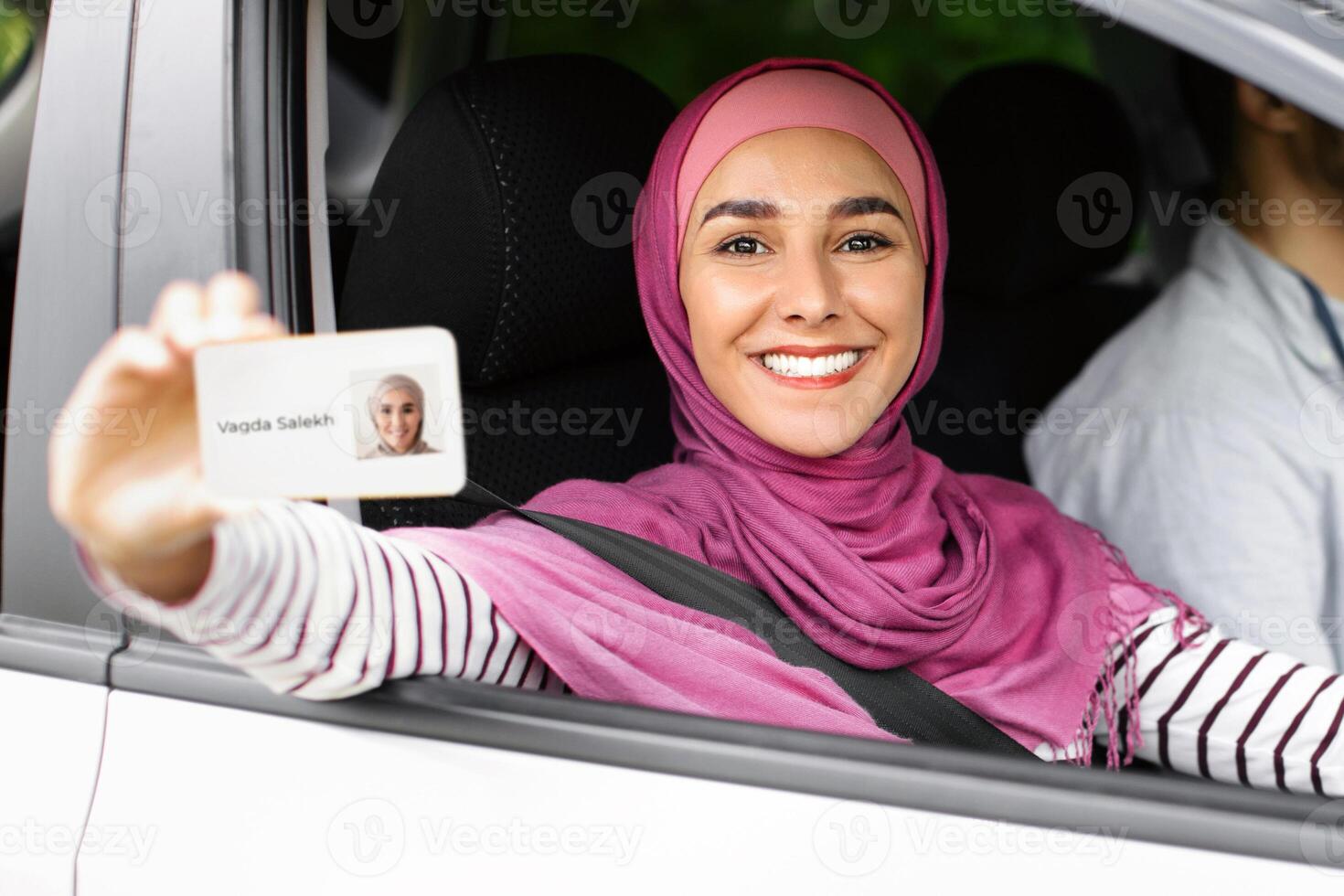 Joyful Islamic Lady In Hijab Demonstrating Driver Licence At Camera While Sitting In Car Next To Instructor, Cheerful Muslim Female In Headscarf Celebrating Passing Exam In Drive School, Closeup photo