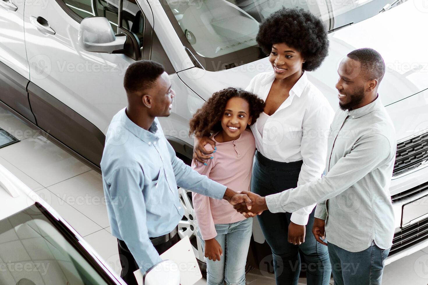 Above view of young black family shaking hands with car salesman, purchasing or renting new vehicle at auto dealership. Manager making agreement with Afro customers at automobile showroom photo