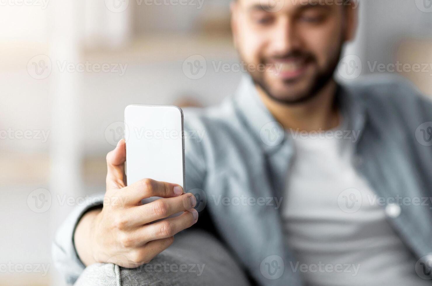 Happy young guy holding smartphone and cup of coffee, sitting on sofa, sending email, browsing web or watching entertainment at home, selective focus. Online life concept photo