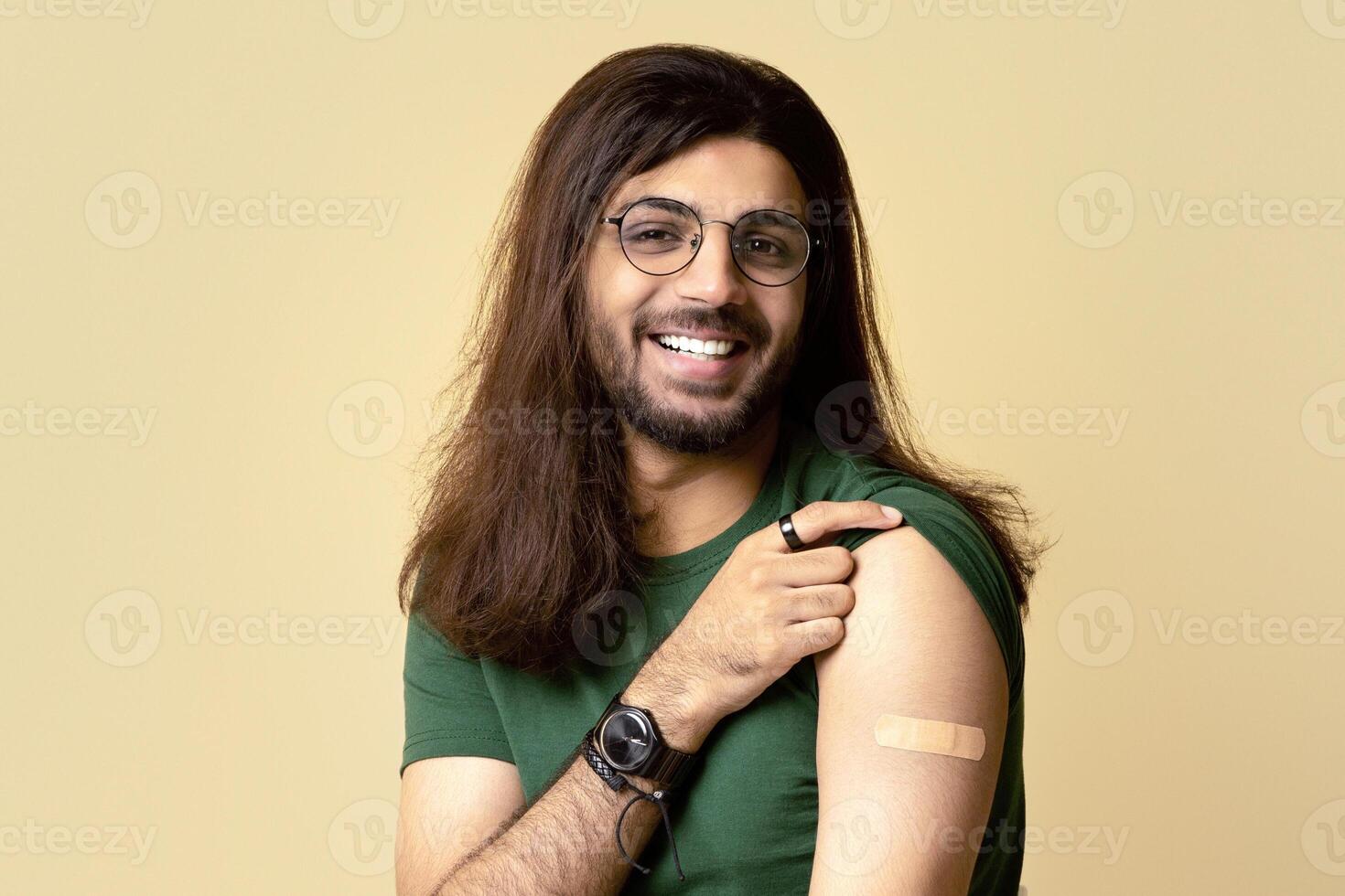 Handsome millennial long-haired arab man got vaccinated against COVID-19, showing shoulder with plaster on and smiling at camera, sitting on chair over beige studio background, copy space photo