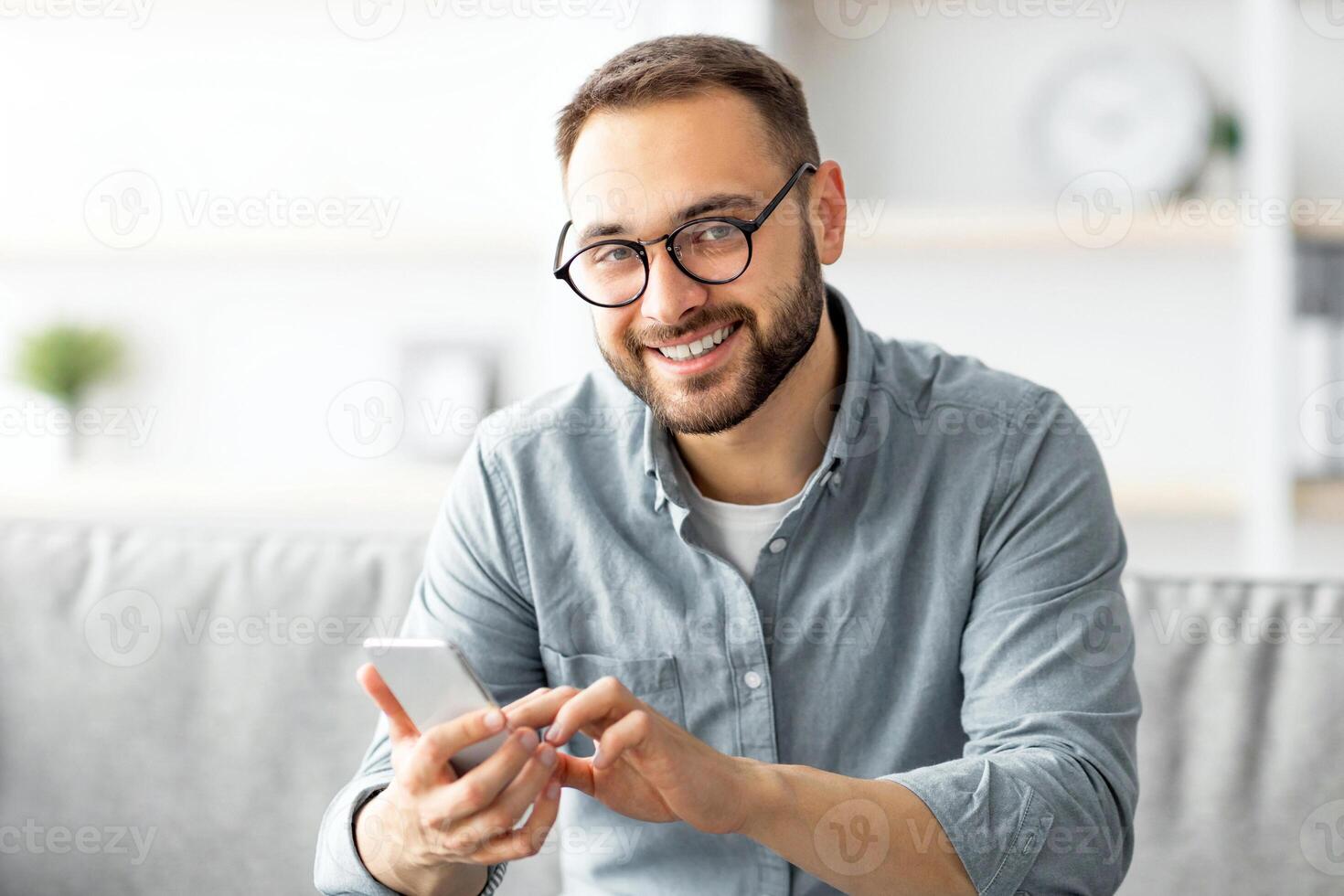 Cheerful millennial guy in glasses sitting on cozy couch with cellphone and messaging to his friend, indoors. Young Caucasian man writing sms, having call or broswing web at home photo