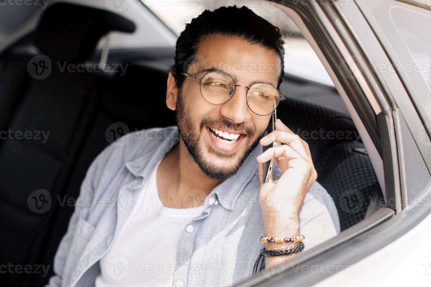 Positive handsome arab guy taking cab, having phone conversation while going to job or home, middle-eastern young man sitting at taxi back seat and talking on smartphone, copy space photo