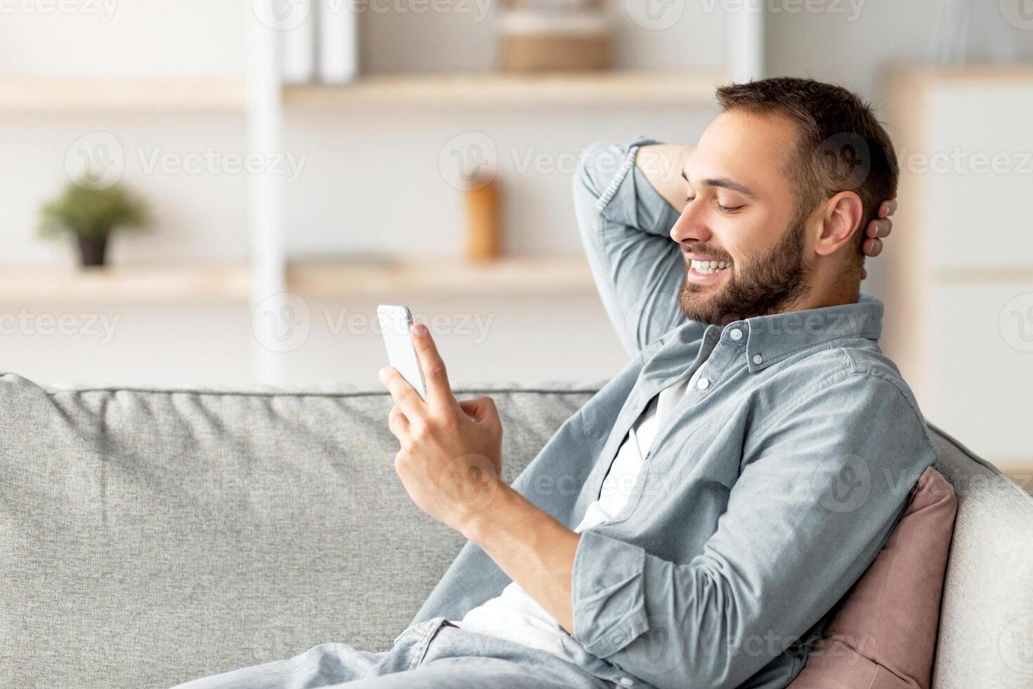 Handsome millennial guy relaxing on sofa with cellphone, smiling and texting his friend at home. Young Caucasian man writing message, having chat or broswing internet at home photo