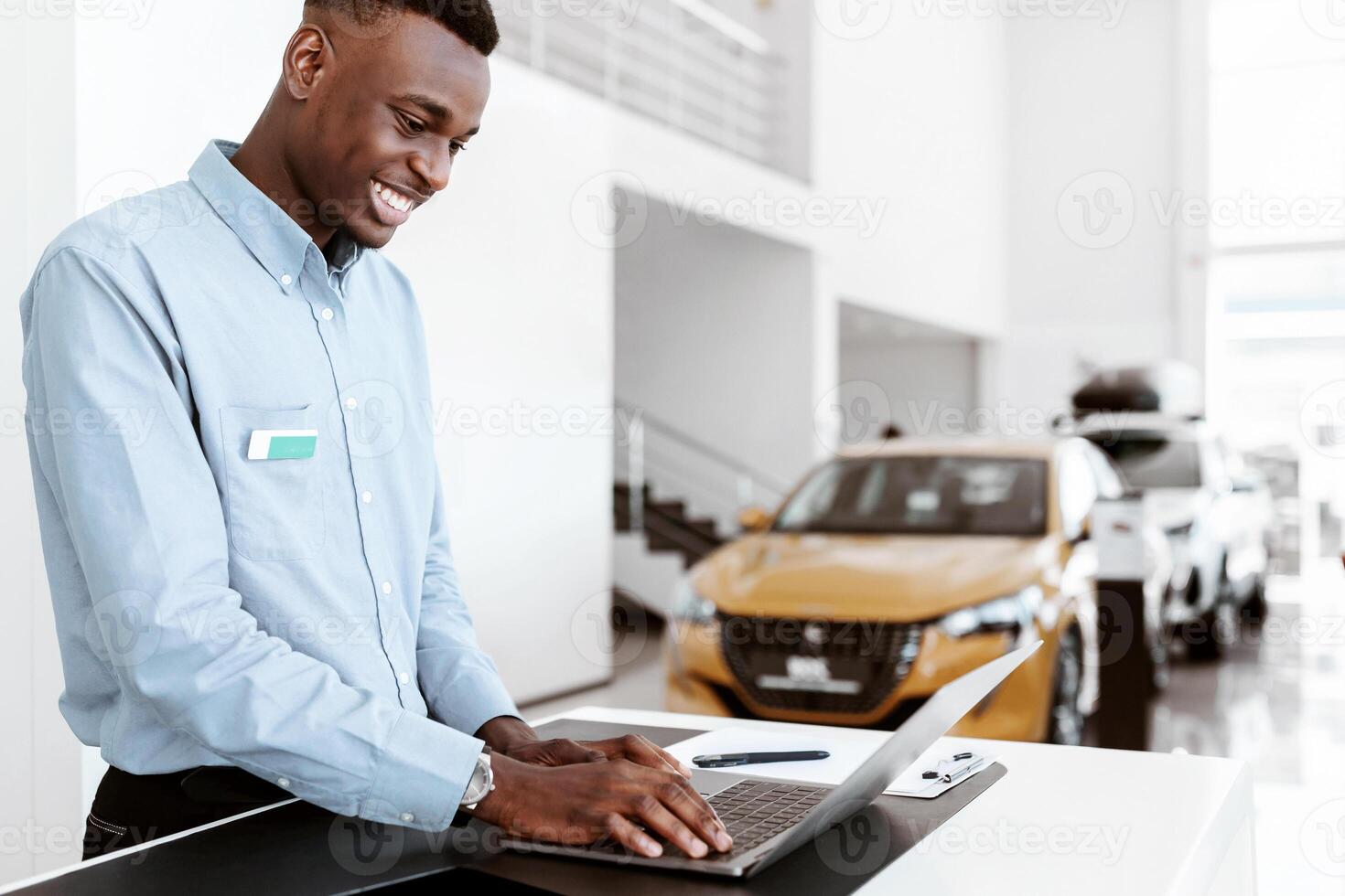 Positive black car salesman using laptop computer at work desk in auto dealership. Cheerful African American automobile manager working with pc in showroom store, free space photo