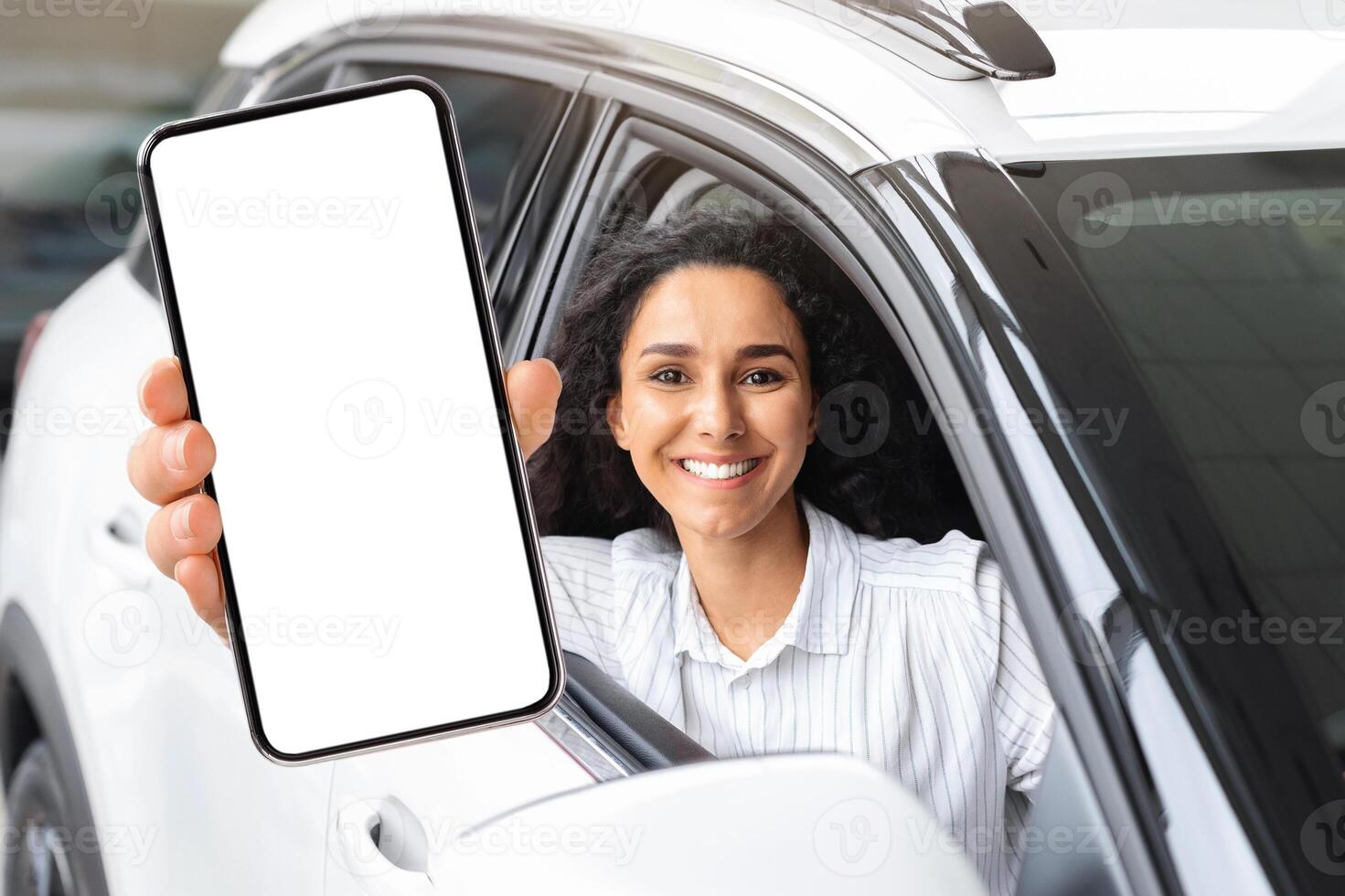 Positive millennial middle eastern woman sitting in brand new white car, showing smartphone with empty screen and smiling at camera, mockup, renting auto on Internet. Car dealership online photo