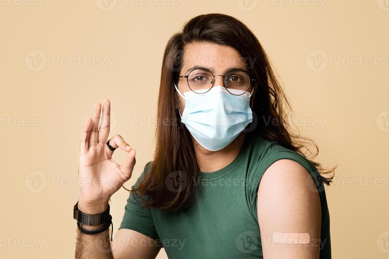 Long-haired arab man in face mask showing okay gesture and plaster on his shoulder, closeup portrait of middle-eastern guy got vaccinated against coronavirus, feeling great, studio background photo