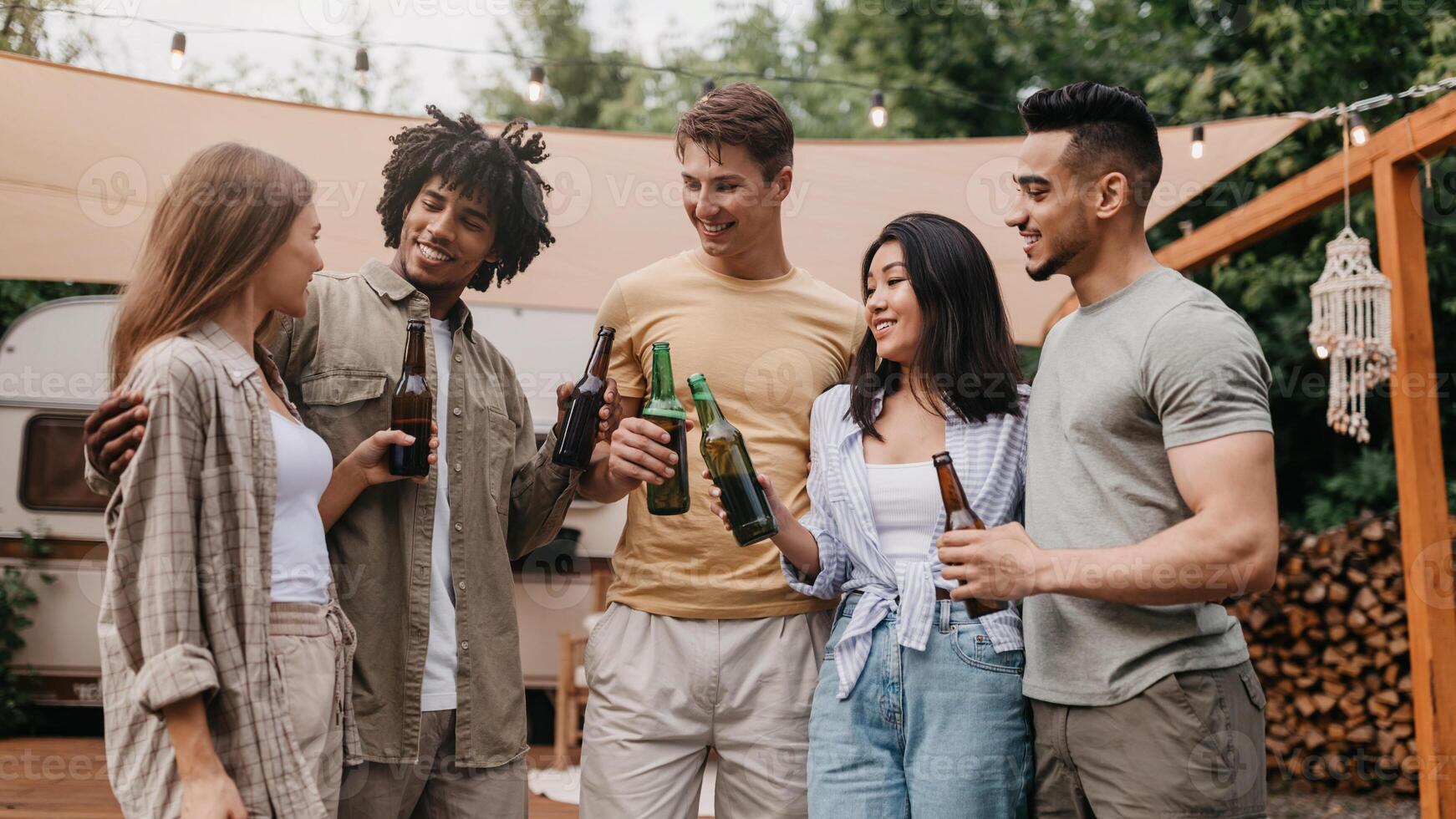 Group of happy multiracial young friends celebrating something, drinking beer, having party near camper van outdoors. Millennial diverse people enjoying summer vacation trip photo