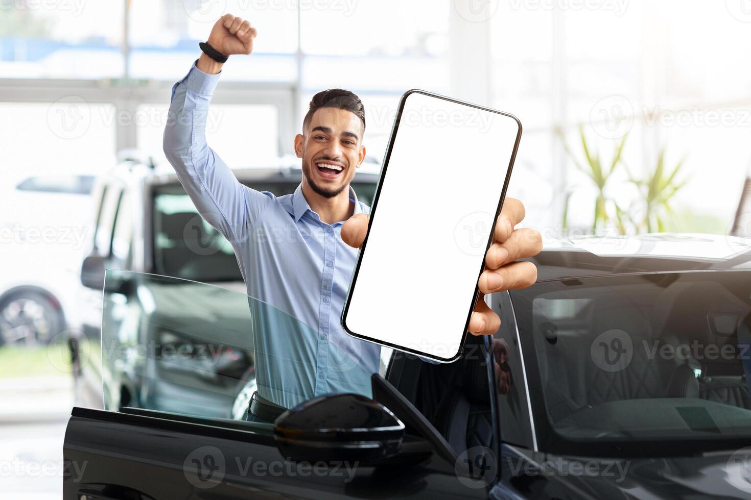 Emotional middle eastern guy choosing new car, standing by brand new black automobile, showing modern cell phone with white blank screen and raissing hand up, using auto dealership app, mockup photo