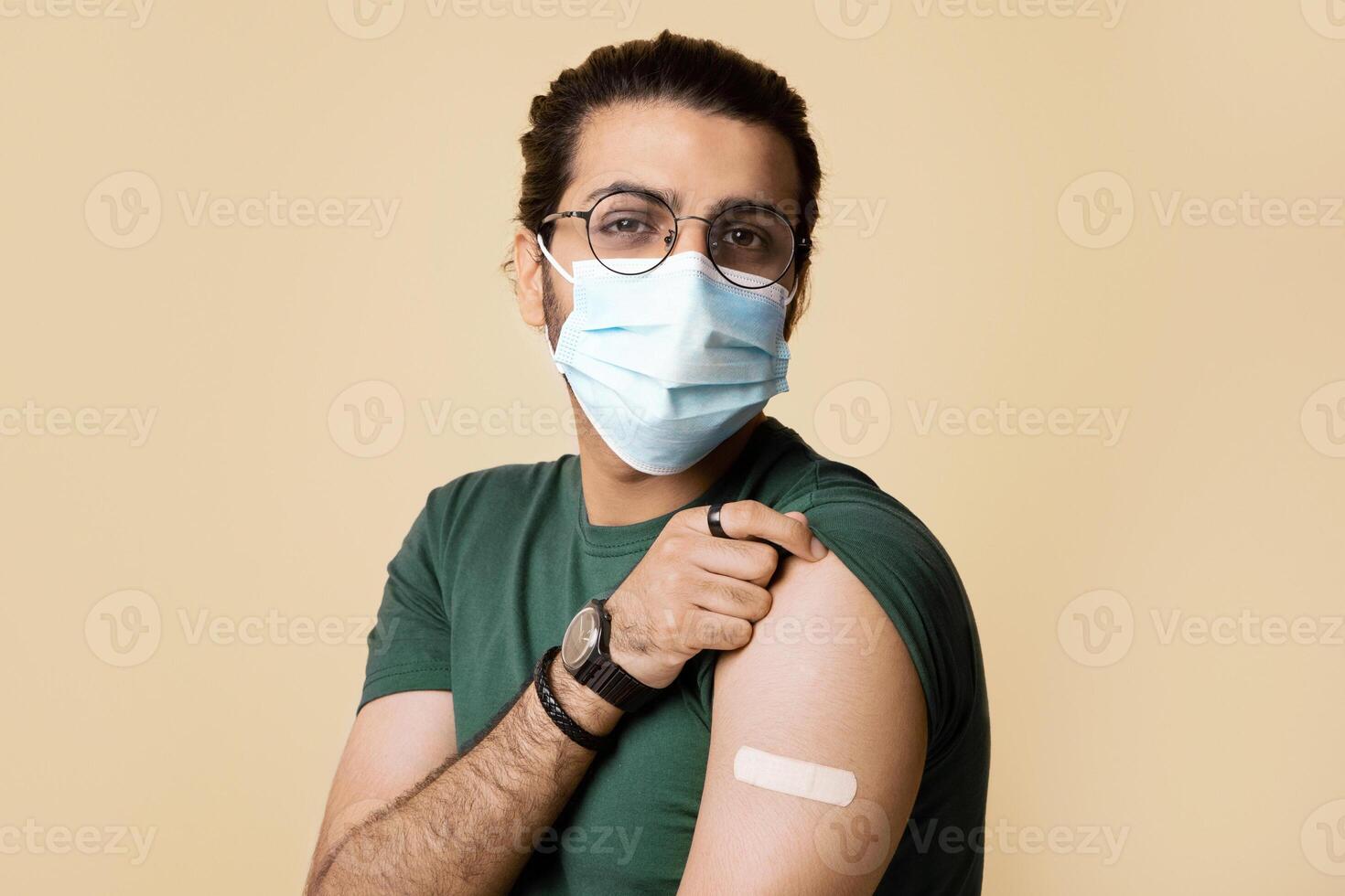 Healthy indian guy in face mask showing medical band on shoulder after vaccination against COVID-19, posing on beige studio background, closeup, copy space. Mass immunization concept photo