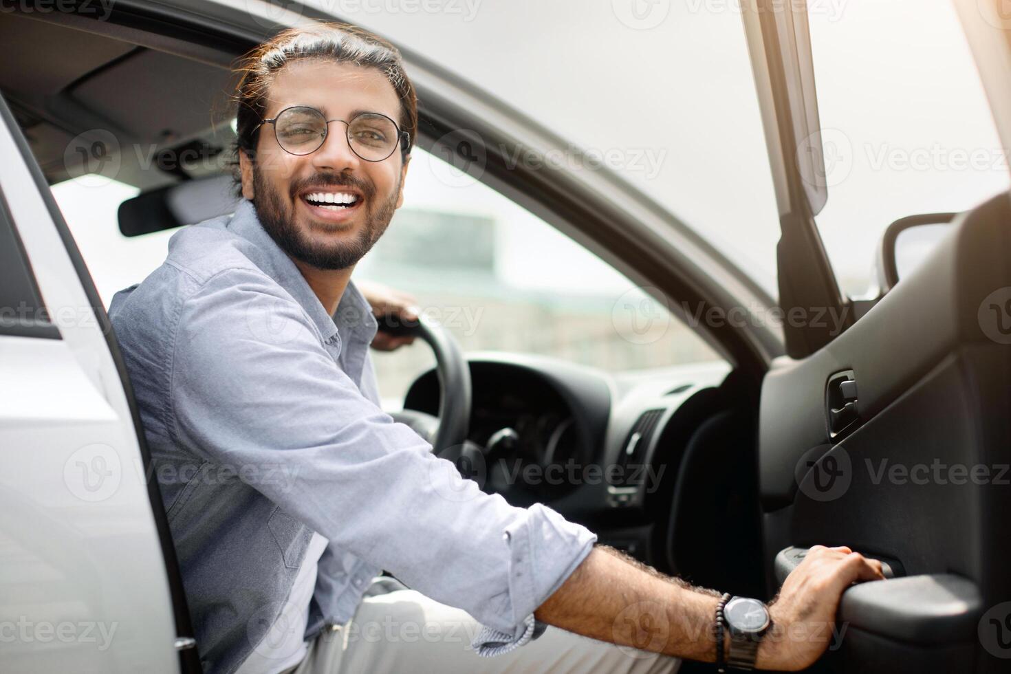 Excited indian guy in casual getting into auto, making test-drive before purchasing, visiting open-air car saloon, smiling at camera while sitting in nice brand new automobile, copy space photo