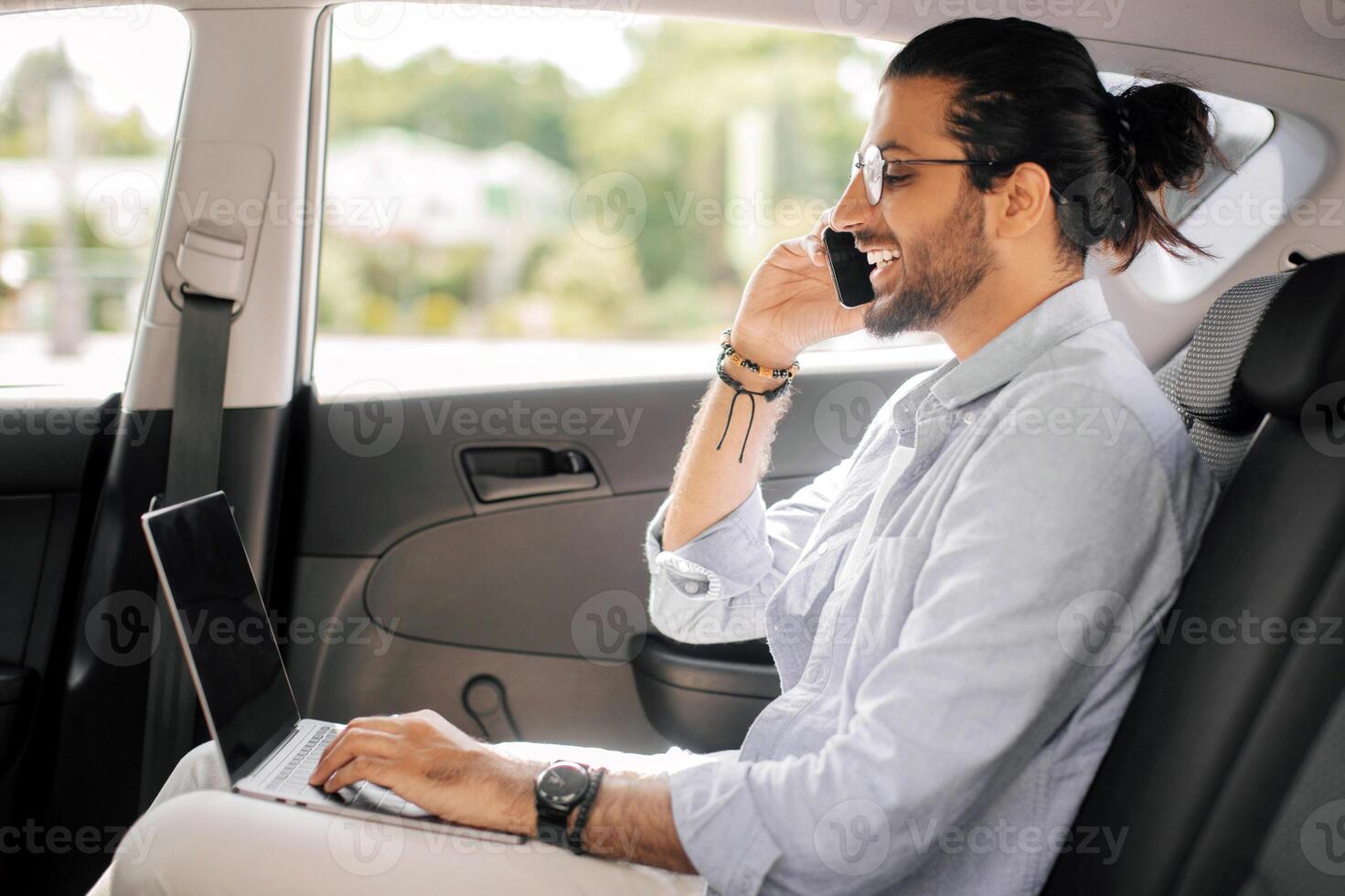 Positive young arab man freelancer working online while sitting at backseat in taxi, typing on laptop with blank screen and having phone conversation, mockup, looking at copy space, side view photo