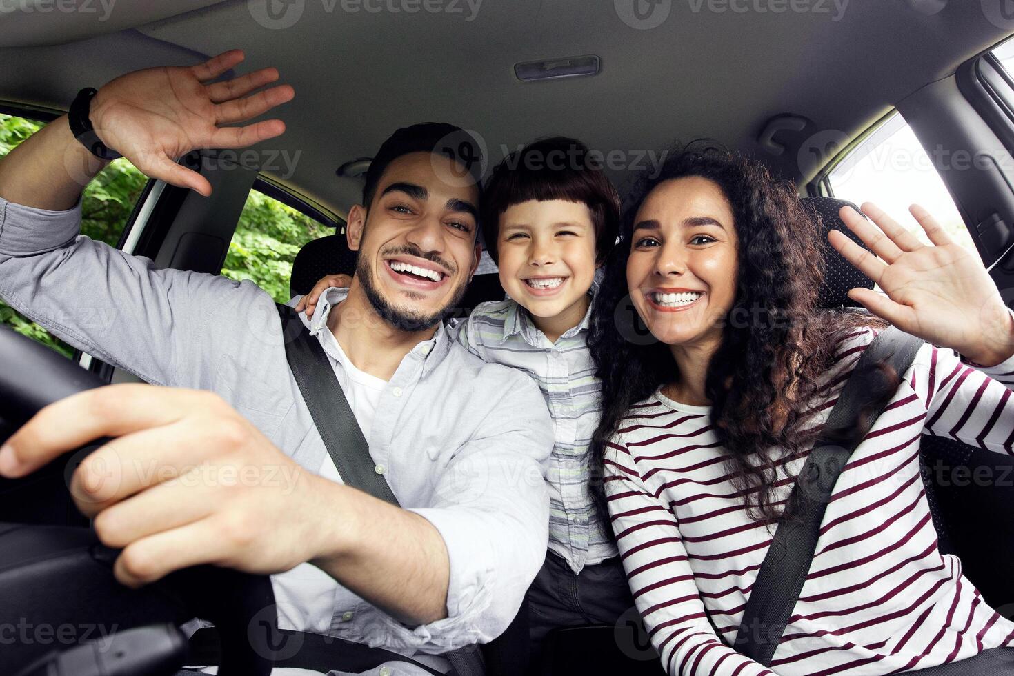 Joyful Middle Eastern Family With Little Son Having Fun While Riding Car Together, Cheerful Arab Parents And Male Child Waving Hands At Camera, Enjoying Road Trip In Their New Vehicle, Closeup Shot photo