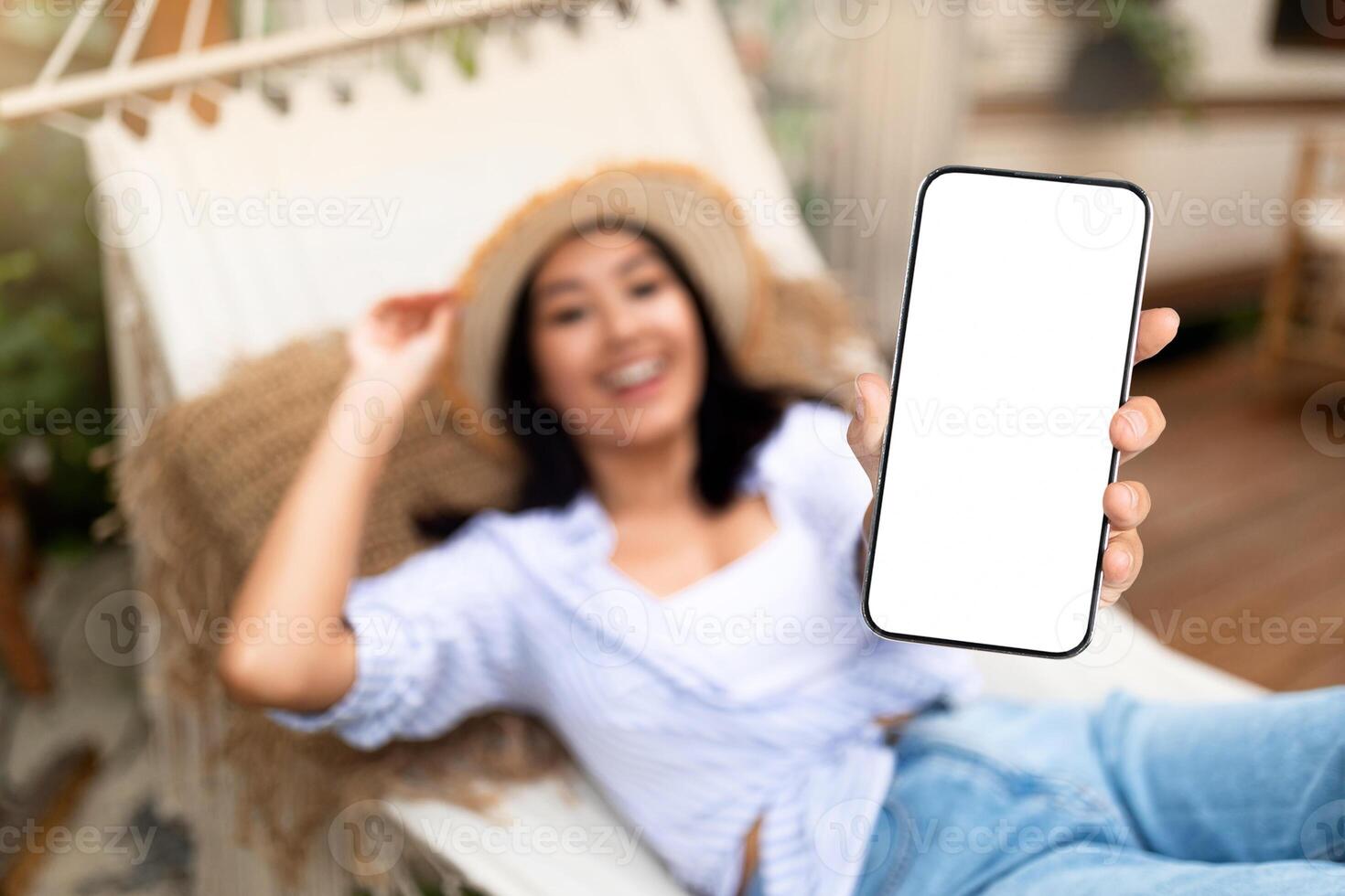 Happy Asian woman lying in hammock near RV, showing smartphone with blank screen on campsite, mockup for mobile app or website design. Selective focus. Cool summer vacation offer concept photo