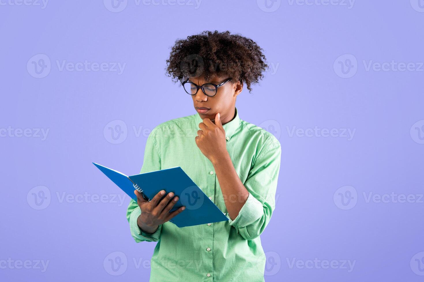 Learning concept. African american male student reading something in notepad, preparing for exams in college, touching chin, standing on blue studio background photo