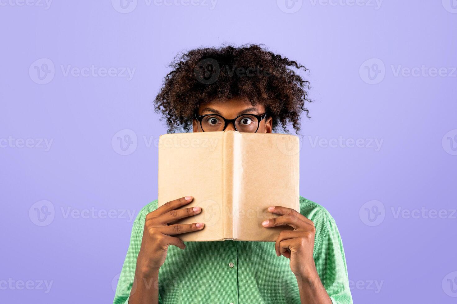 University exams. Shocked african american guy in glasses covering face with book, hiding behind it with eyes wide open, standing over blue studio background photo