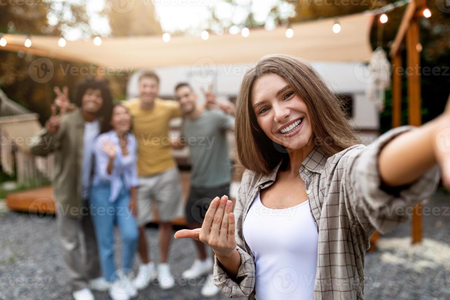 Lovely Caucasian woman taking selfie with her multiethnic friends next to motorhome on campsite, free space. Pretty young lady making mobile photo on autumn camping trip