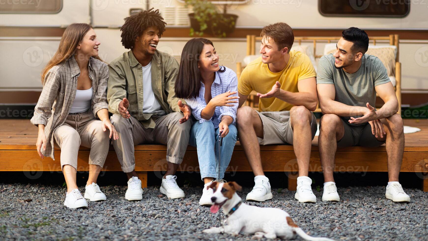 Positive diverse friends and their pet dog sitting near motorhome, having fun together outdoors. Group of multiracial millennials relaxing on camping trip, speaking to each other in countryside photo