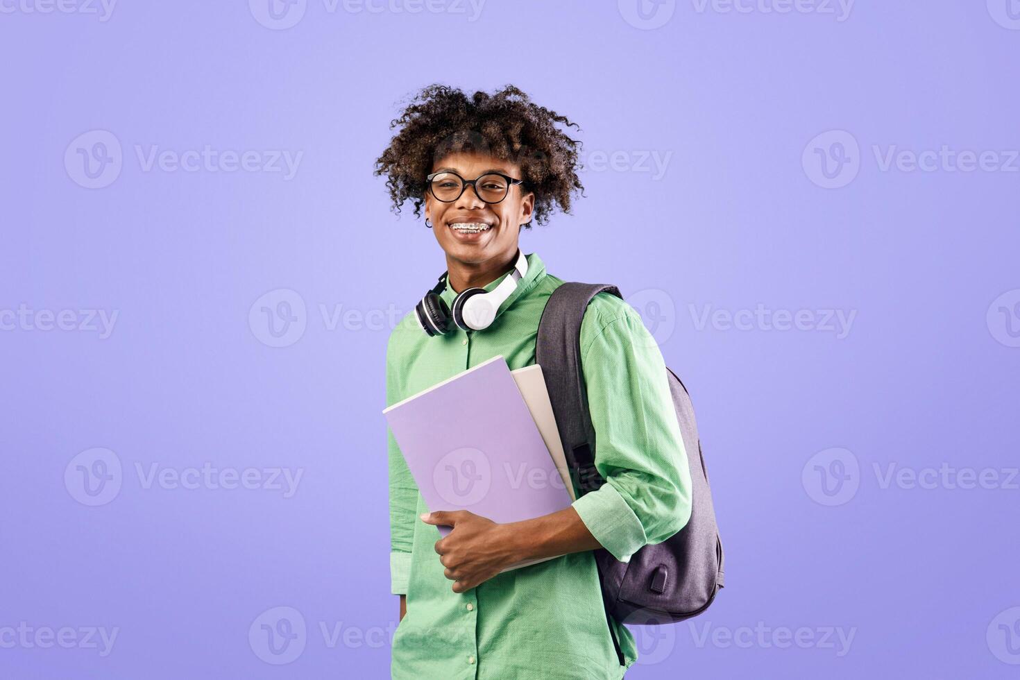 Smart african american student guy with backpack and notebooks smiling at camera, posing over blue studio background. Education, university, college, studying, course concept photo