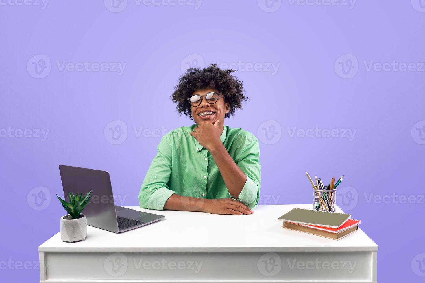 Online school or educational course concept. Excited smiling black guy in eyeglasses sitting at the desk and using laptop, doing homework, posing to camera over blue background photo
