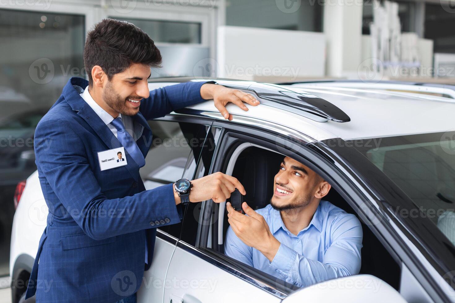Cheerful sales manager handsome middle eastern man in blue suit giving car key to owner sitting inside white automobile, happy arab guy buying new auto at dealersip salon, copy space photo