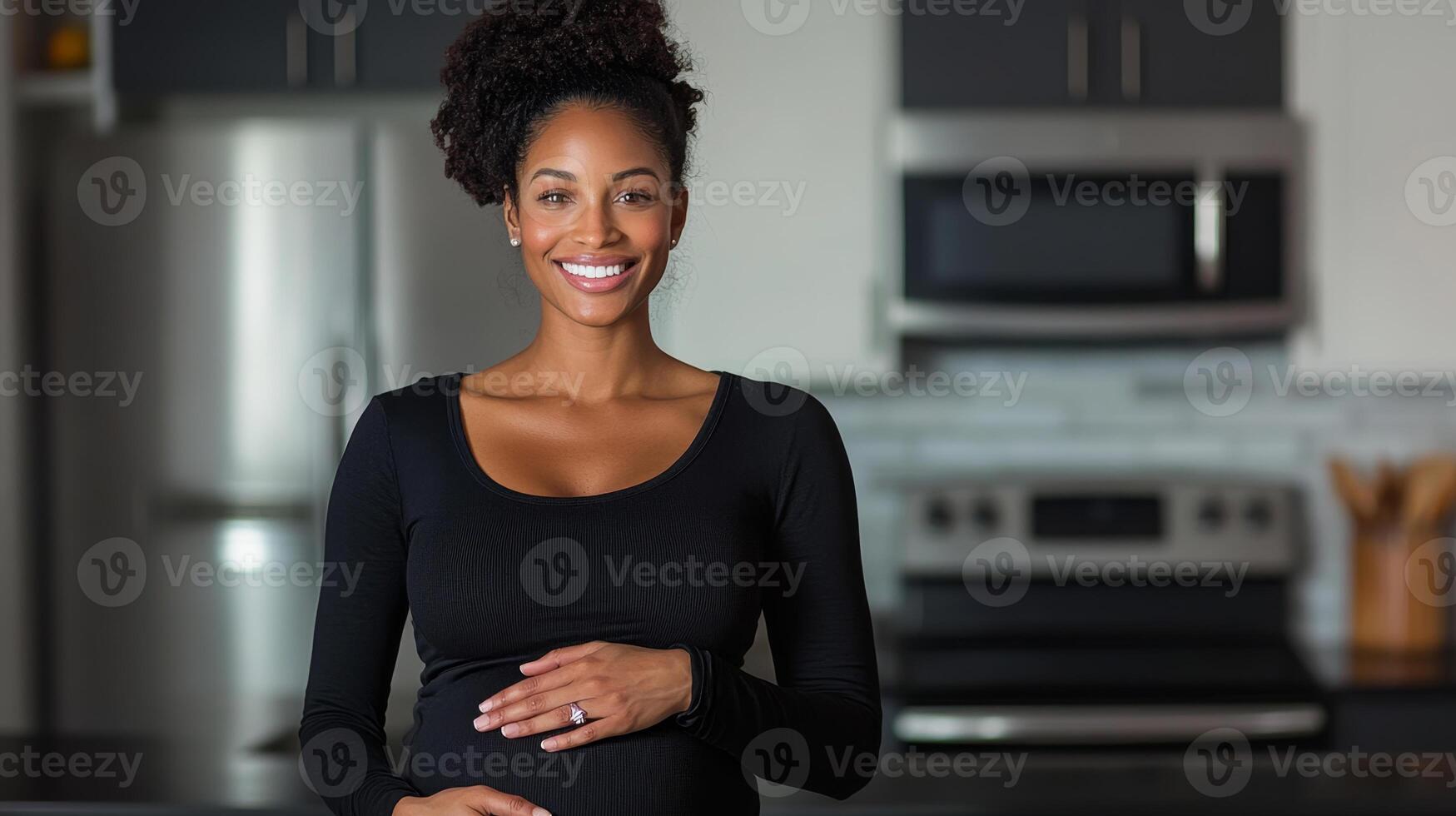 A pregnant woman stands in a contemporary kitchen, gently holding her belly with a joyful smile. The bright space features stainless steel appliances, reflecting a warm and welcoming atmosphere. photo