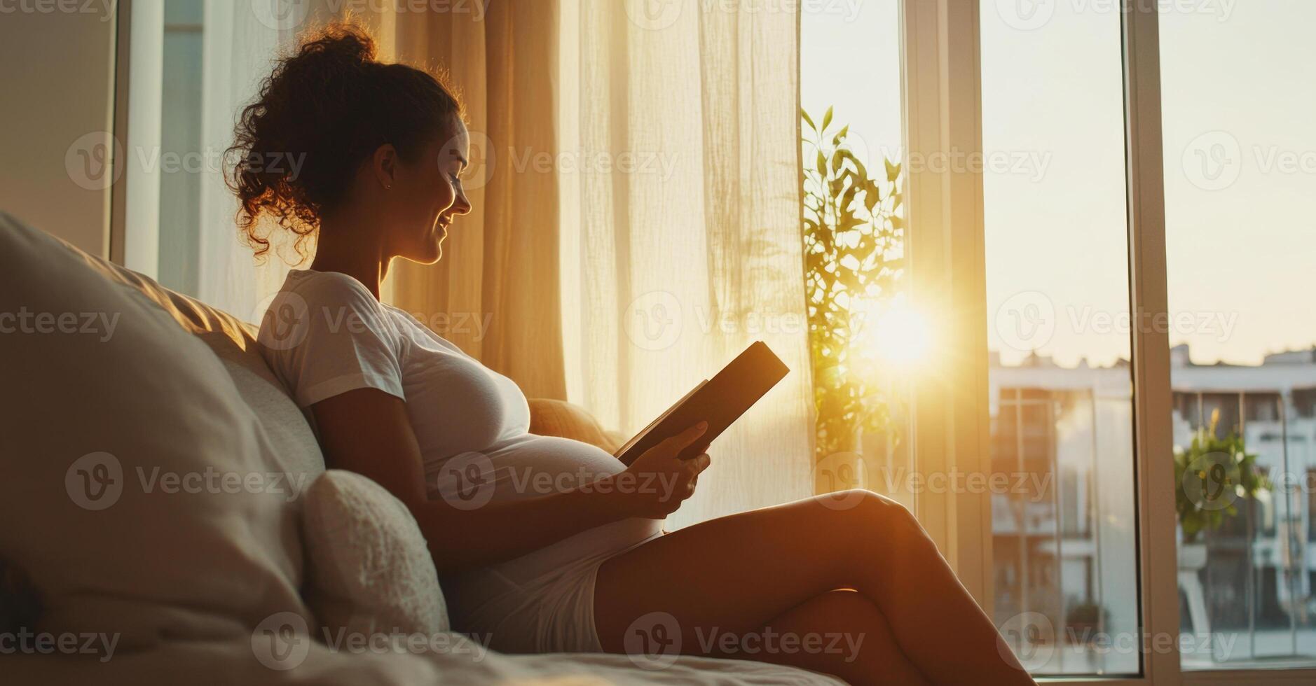 A pregnant woman sits comfortably on a sofa, enjoying a book as the warm sunlight streams through the window, creating a serene and cozy atmosphere at home during sunset. photo