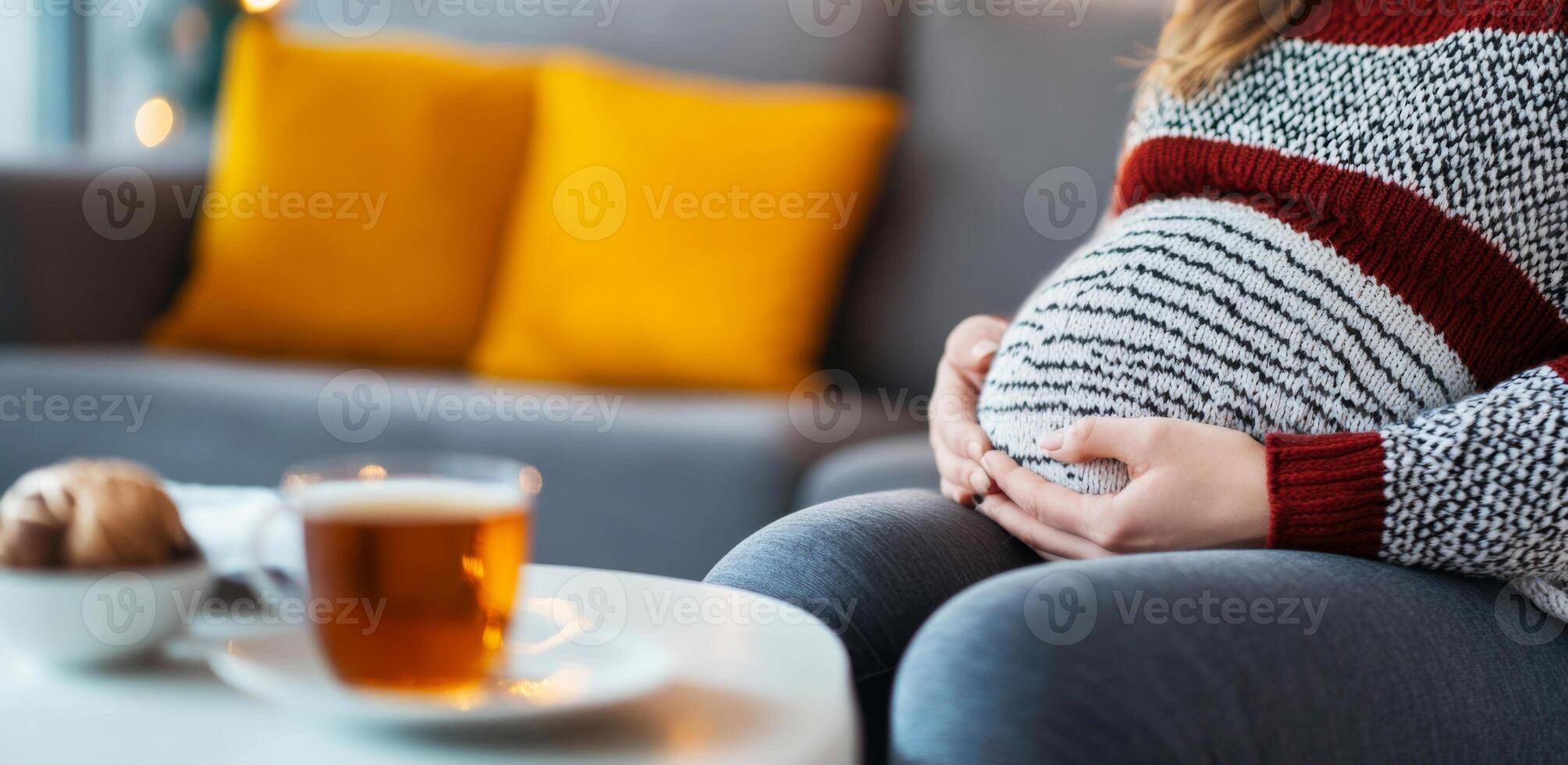 A pregnant woman sits comfortably at home, cradling her baby bump while sipping tea. Soft pillows decorate the couch, creating a warm and inviting atmosphere for relaxation. photo