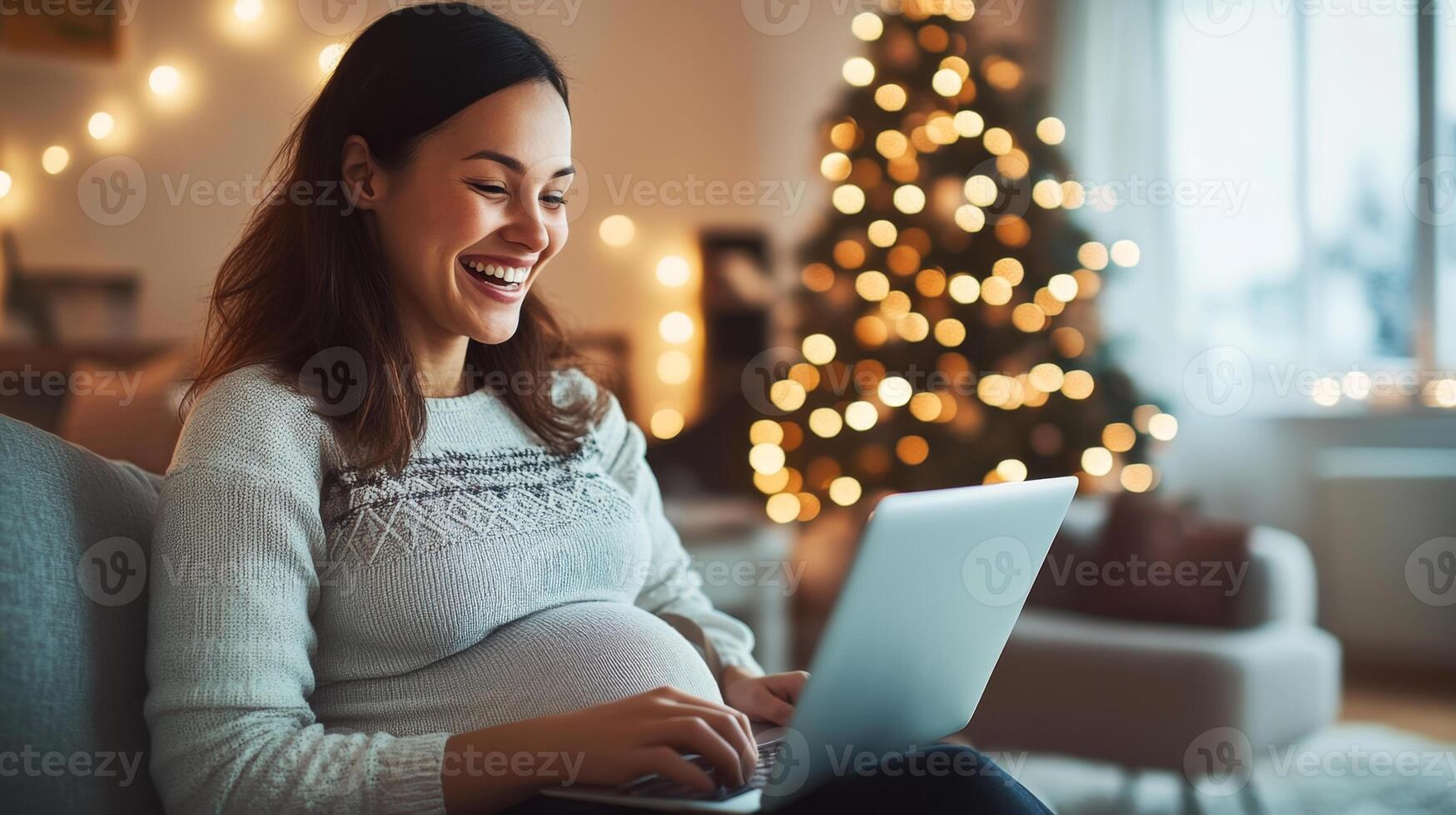 A woman in late pregnancy relaxes at home, smiling as she uses her laptop. Warm lights and a decorated Christmas tree create a cozy atmosphere, enhancing the peaceful moment of joy and comfort. photo
