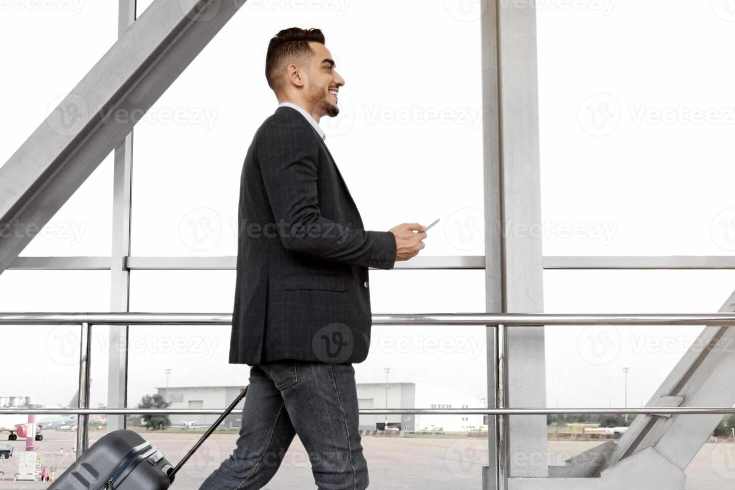Young middle-eastern businessman with suitcase and smartphone in hands walking in airport terminal, handsome arab male going to flight boarding, ready for air trip, side view with copy space photo