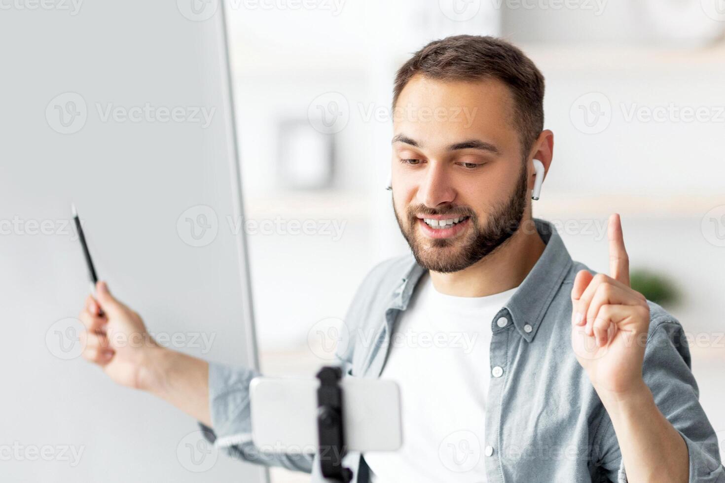 Happy male teacher giving online private lesson from home, pointing at empty blackboard with empty space. Millennial tutor communicating with pupils on webcam, using cellphone photo