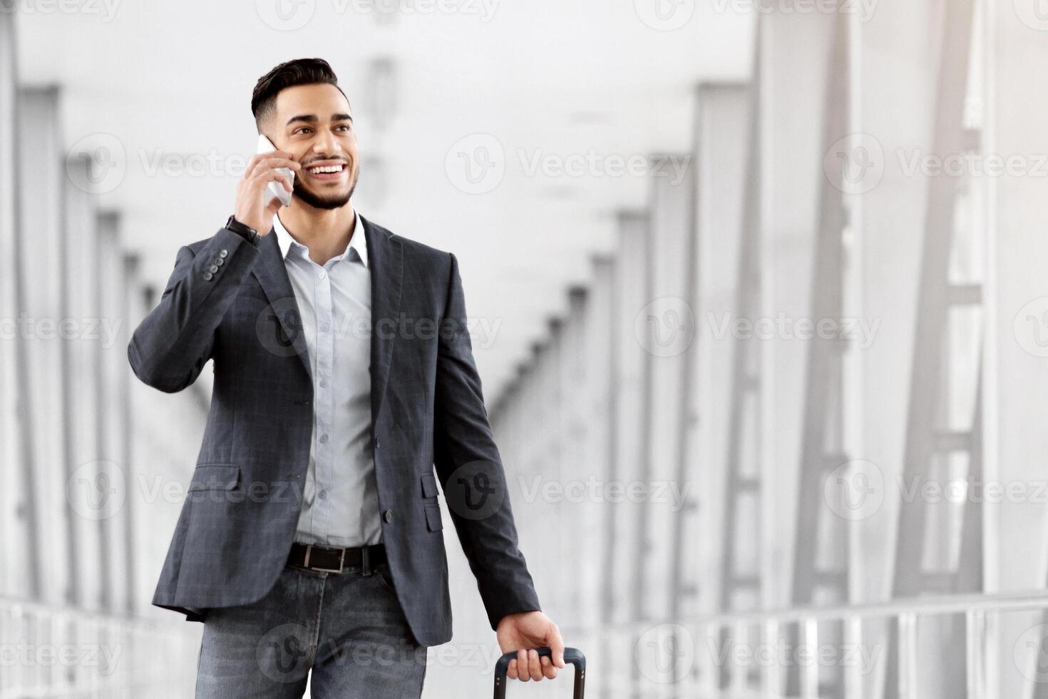 Handsome Arab Man Walking With Suitcase In Airport And Talking On Cellphone, Young Middle Eastern Businessman Booking Transfer By Phone After Arrival, Enjoying Pleasant Conversation, Copy Space photo
