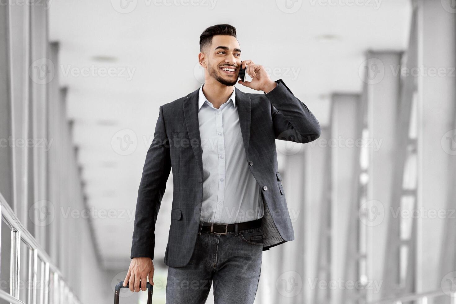Handsome young arab businessman talking on cellphone and carrying suitcase in airport, smiling middle eastern man walking with luggage to flight gate and enjoying mobile conversation, copy space photo