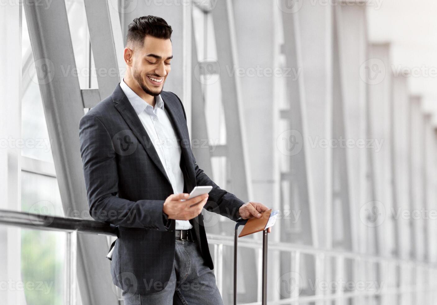 Handsome Arab Man Relaxing With Smartphone While Waiting For Flight In Airport, Smiling Middle Eastern Male Traveller Using Mobile Phone In Terminal, Booking Hotel Online Or Browsing Internet photo