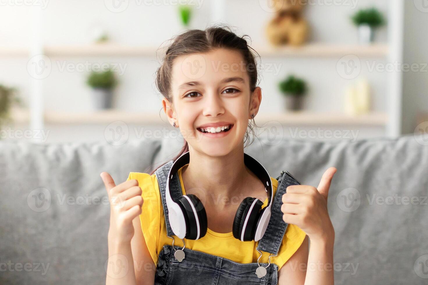 Distant Education. Portrait Of Cheerful Adolescent Girl With Headphones Using Laptop In Living Room Sitting At Table And Showing Thumb Up Gesture Looking At Camera, Pupil Studying Online With Computer photo