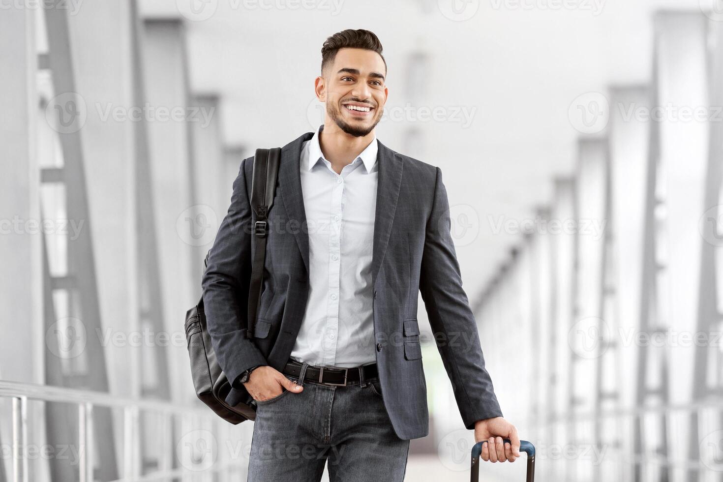 Portrait Of Handsome Happy Arab Man Walking In Airport Terminal With Suitcase, Smiling Young Middle Eastern Guy In Casual Clothes Going To Boarding Gate, Enjoying Upcoming Air Trip, Copy Space photo
